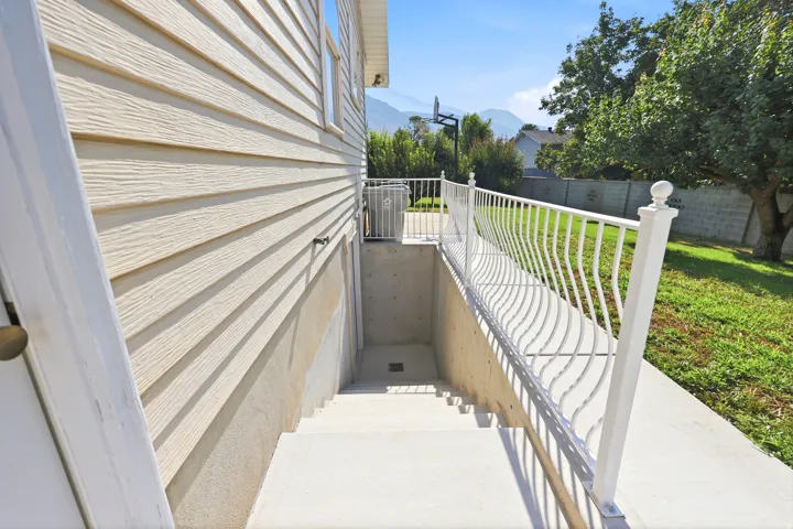 Concrete basement entrance with white metal railing, textured horizontal siding, and a partial backyard with mature trees