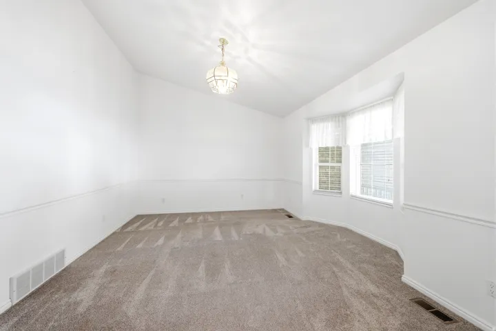 Bright room featuring a vaulted ceiling with a decorative chandelier, a bay window with horizontal blinds and valances, and light-toned carpet flooring