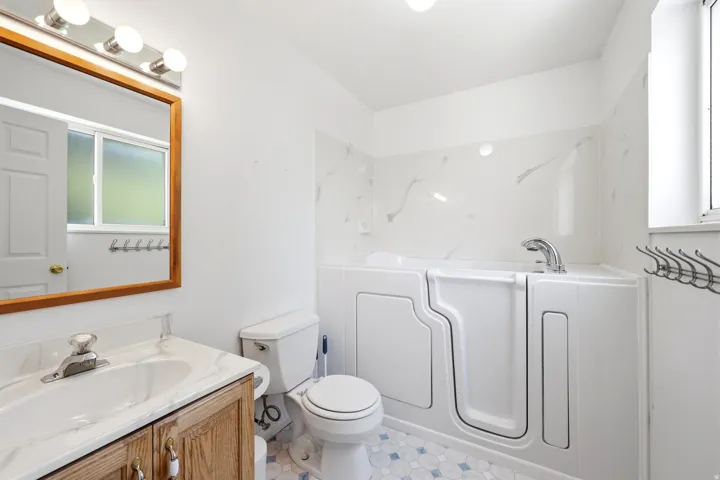 Bathroom featuring a walk-in tub, white vanity with wood-finish cabinetry, a single basin sink, and a framed mirror with overhead lighting