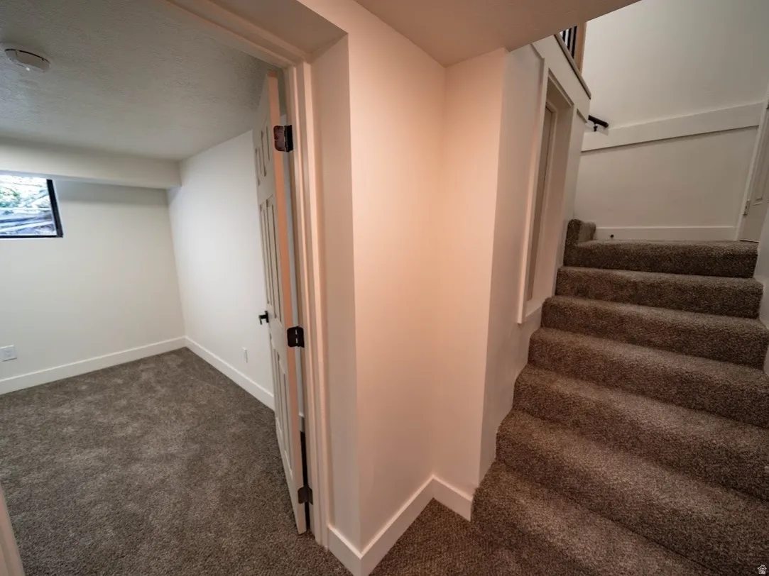 Carpeted stairway with a black metal handrail leading to a lower level featuring a room with gray carpet, white walls, and a single window