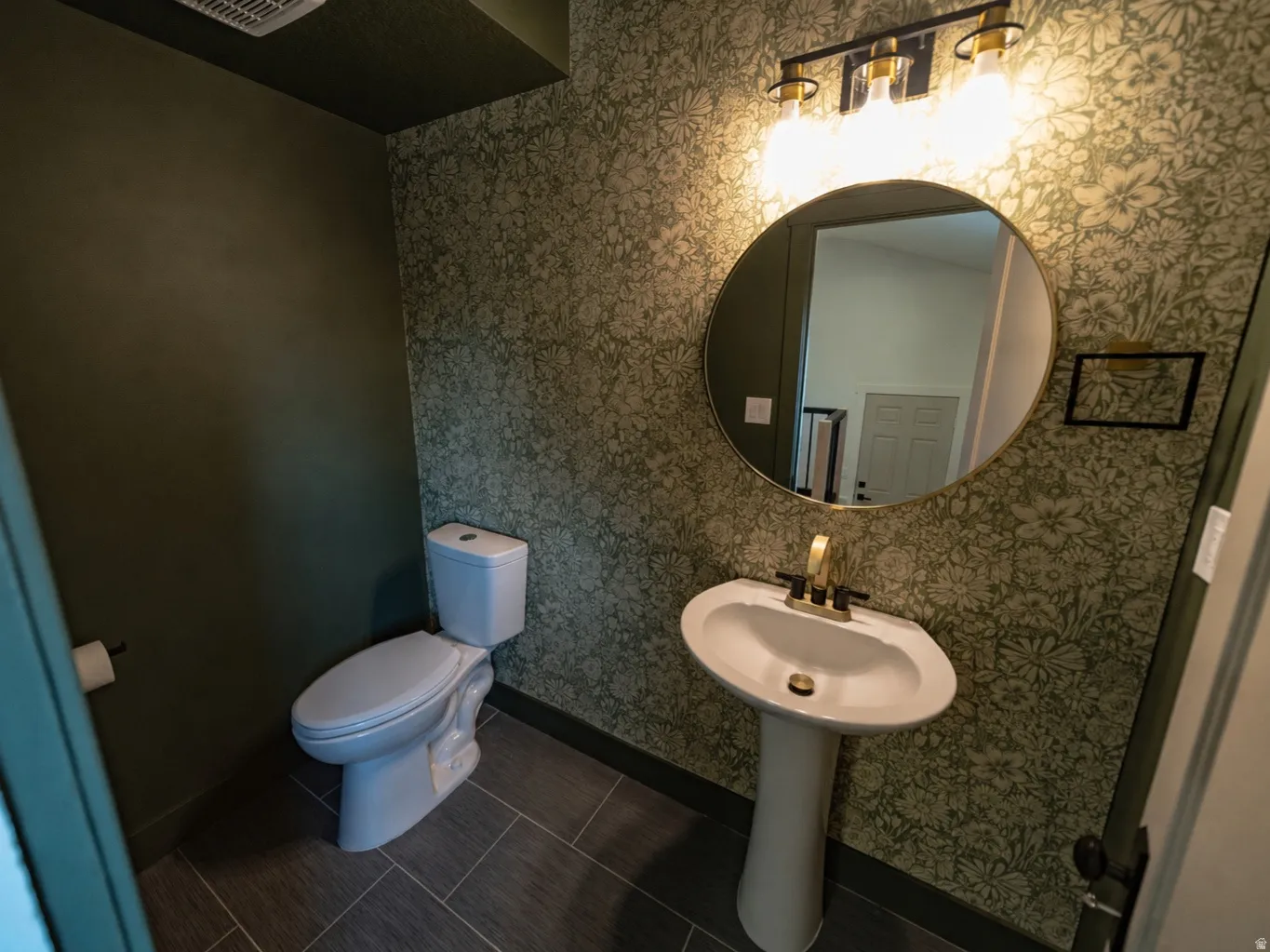 Half Bath with pedestal sink with gold-tone faucet, round mirror, three-light vanity fixture, patterned wallpaper, and rectangular floor tiles