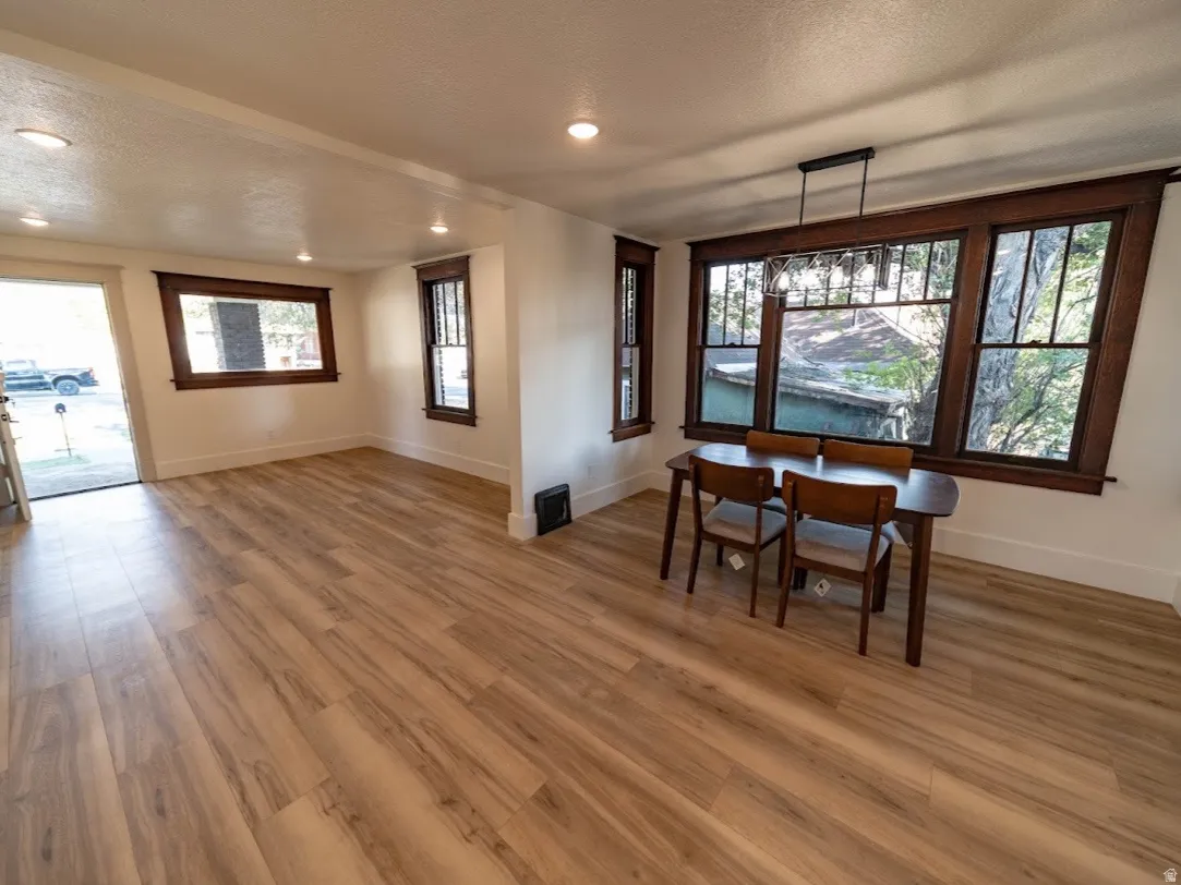 Spacious open-concept living area showcasing warm wood-finish flooring throughout, abundant natural light from multiple large windows and glass doors, recessed ceiling lighting, and a dedicated dining zone with built-in seating area