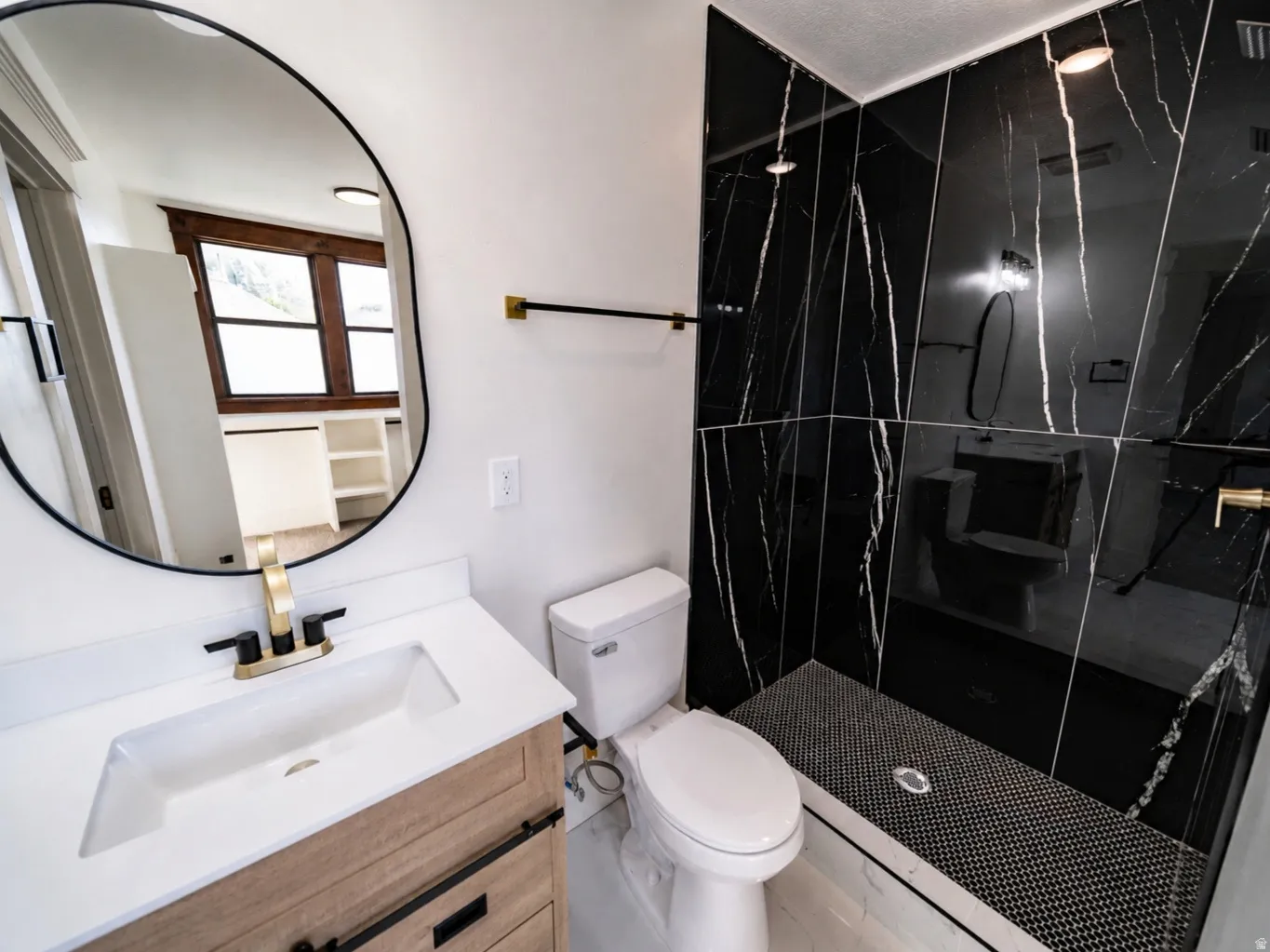 Bathroom featuring a wood-finish vanity with an integrated sink, a frameless oval mirror, a walk-in shower with black-veined wall tiles, black mosaic floor tiles, and gold-finish fixtures