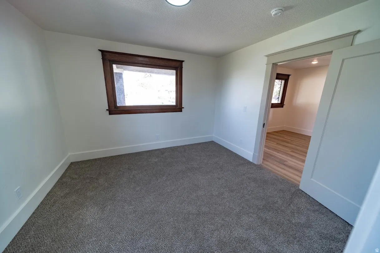 Bright bedroom featuring neutral wall finishes, carpeted flooring, a large casement window with wood trim, and direct access to adjoining spaces through wide doorways. Second bedroom on main floor.