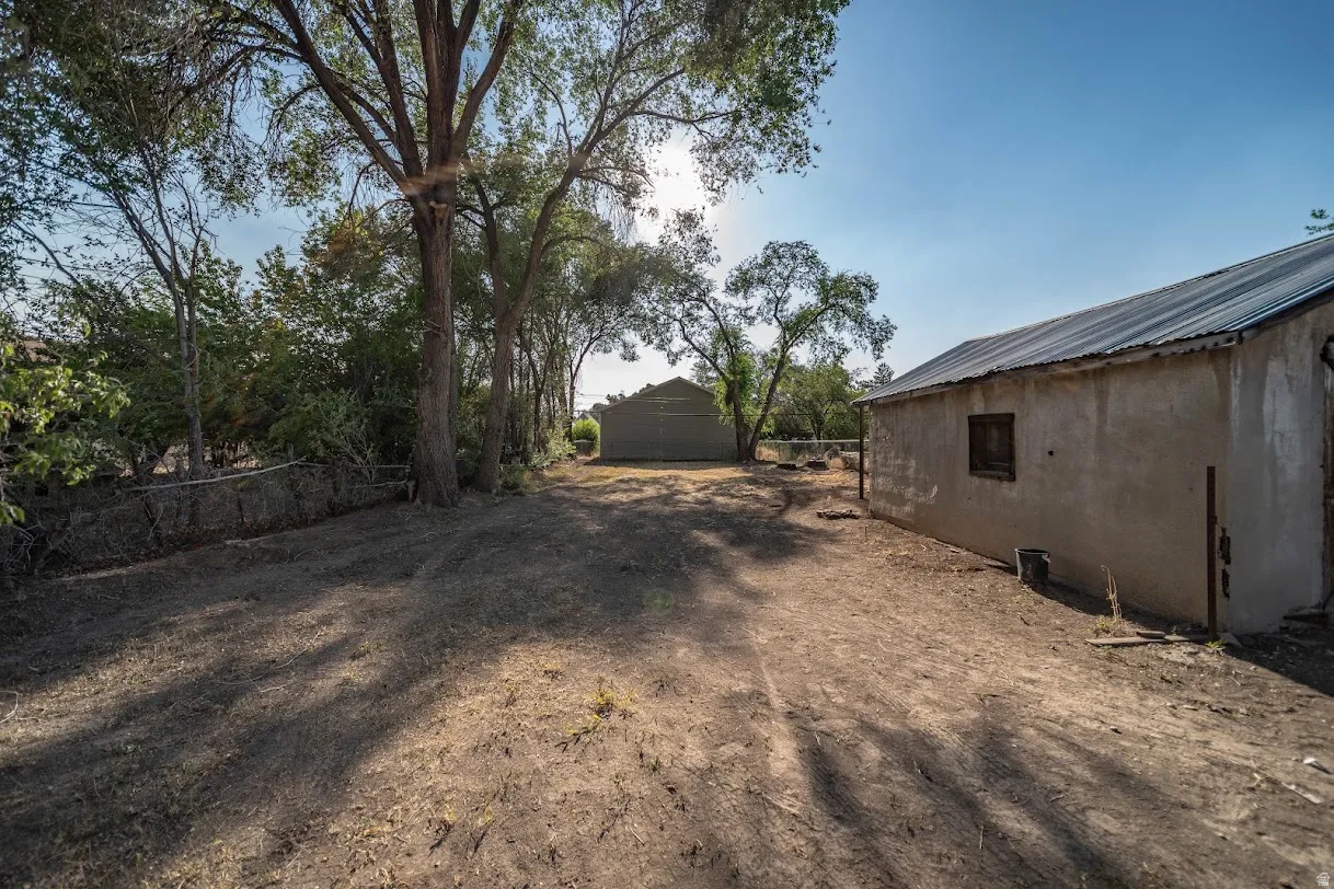 Expansive outdoor space featuring a stucco-finish building with a corrugated metal roof, mature trees providing natural shade, and a secondary structure in the background