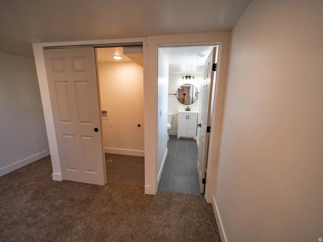 Carpeted room with white walls, featuring a laundry alcove with tile flooring and a panel door, and a bathroom entry with a white vanity, circular mirror, and tile flooring