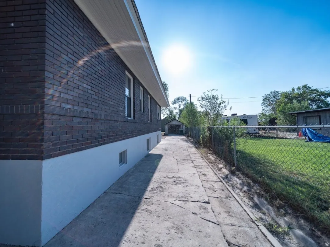 Brick exterior with white foundation trim, concrete pathway, chain link fencing, and a detached structure in the background