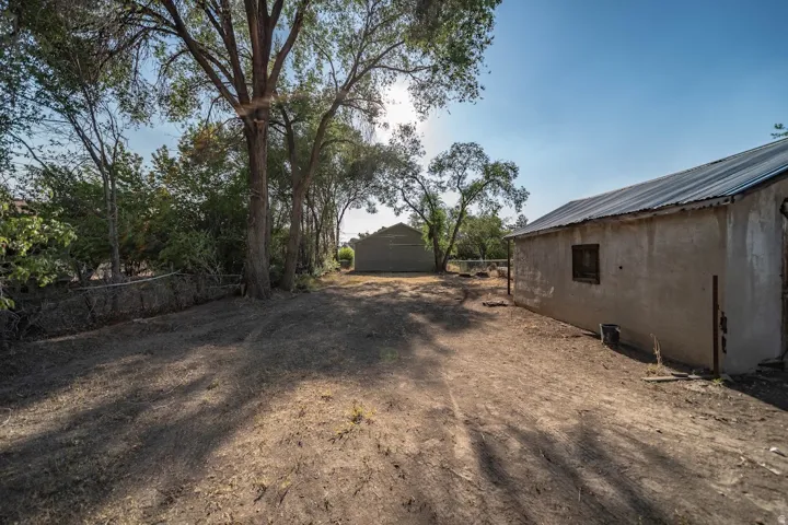 Expansive outdoor space featuring a stucco-finish building with a corrugated metal roof, mature trees providing natural shade, and a secondary structure in the background