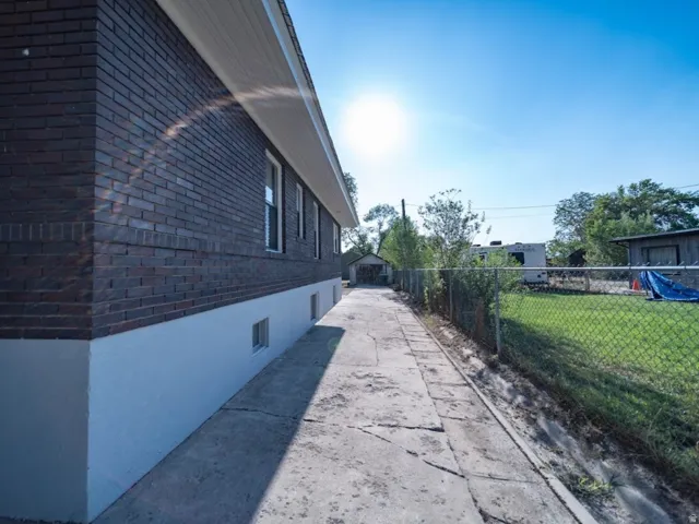 Brick exterior with white foundation trim, concrete pathway, chain link fencing, and a detached structure in the background