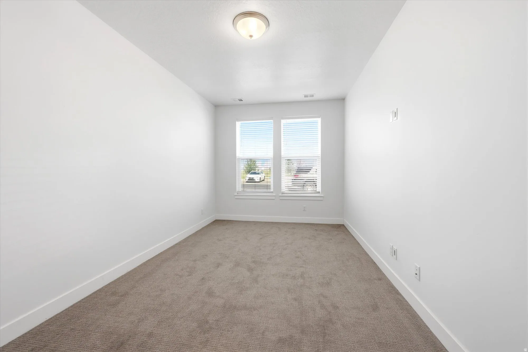 Carpeted interior room featuring two windows with horizontal blinds, white walls, and a ceiling-mounted light fixture