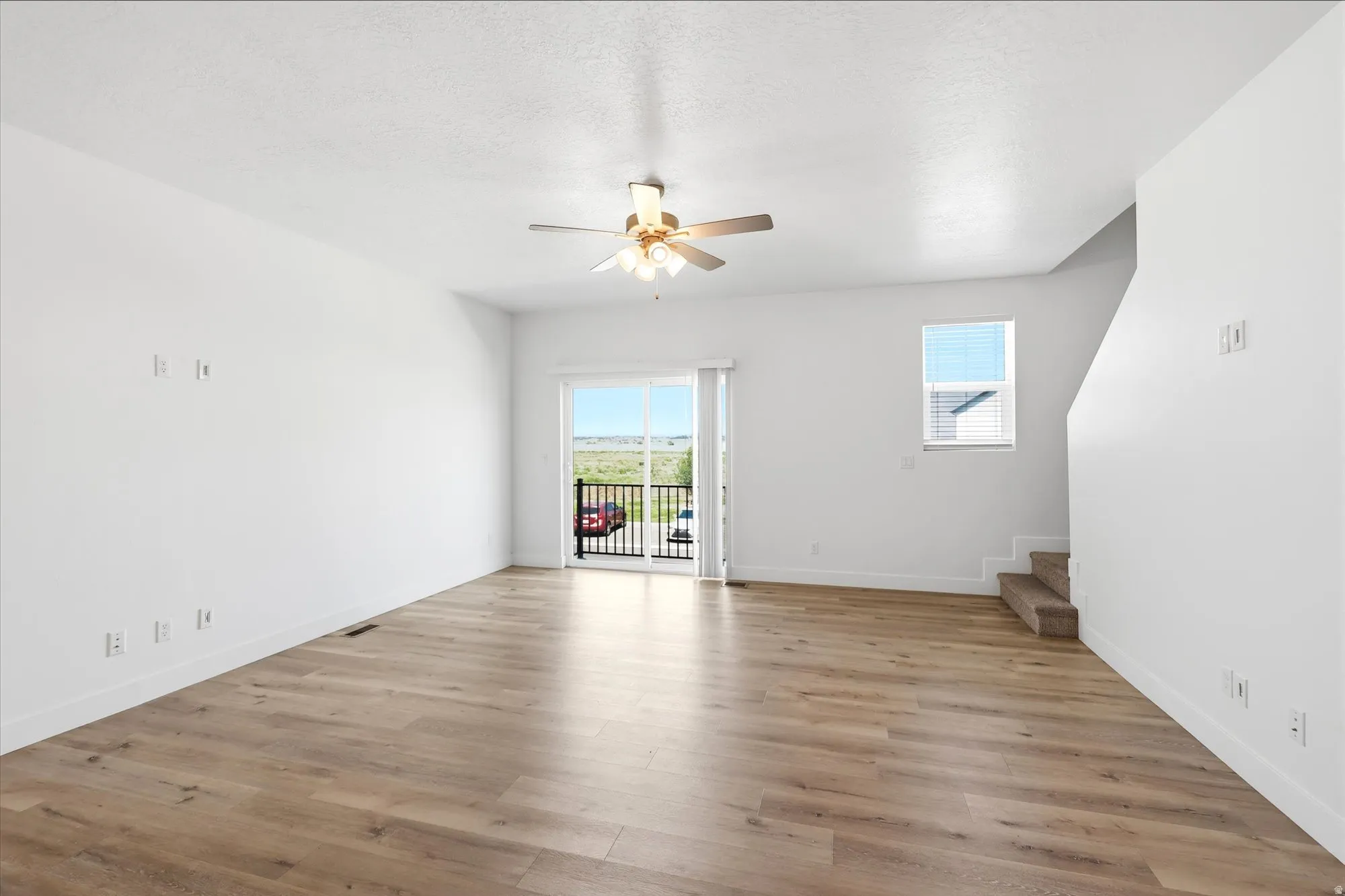 Spacious living area featuring wood-finish flooring, a ceiling fan with integrated lighting, a sliding glass door opening to a balcony, and a window with horizontal blinds