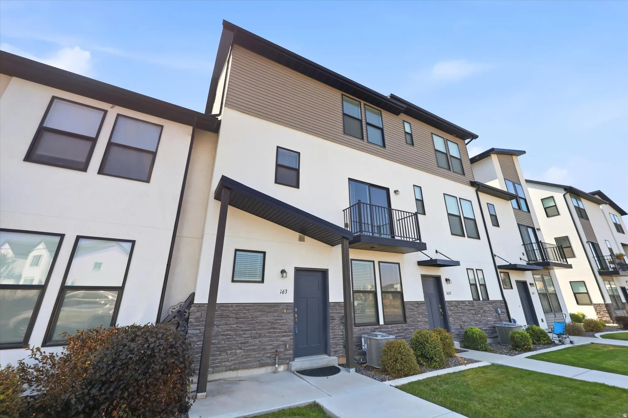 Modern townhome exterior featuring a multi-level facade, dark-framed windows, a black metal balcony, a covered entryway, and stone veneer accents