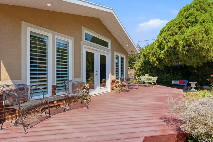 Expansive outdoor deck with red-toned wood-finish flooring, stucco exterior, multiple windows with plantation shutters, and double French doors