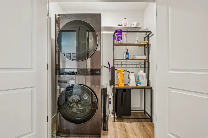 Dedicated laundry closet featuring stacked front-load washer and dryer with glass doors, wood-finish flooring, and industrial-style metal shelving with multiple tiers for storage of cleaning supplies and household essentials