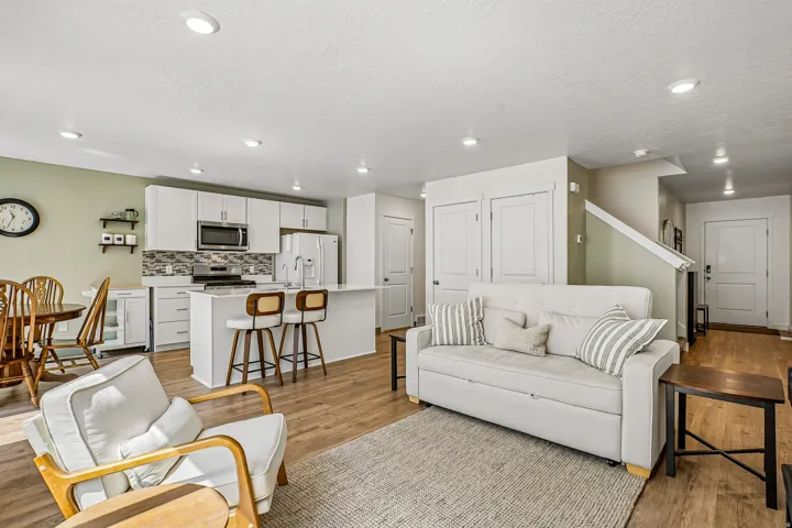 Open-concept living area featuring a white cabinetry kitchen with stainless steel appliances and a centered island with seating, wood-finish flooring throughout, recessed ceiling lighting, a cream upholstered sofa, natural wood accent chairs, and neutral