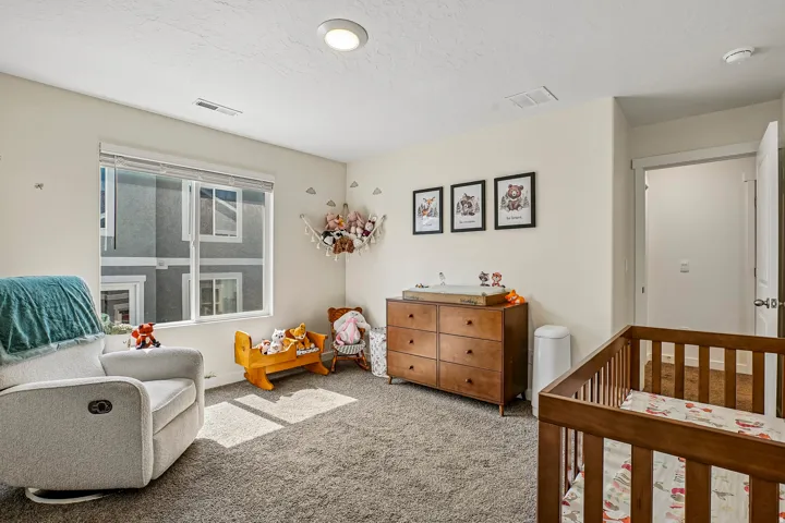 Bright nursery featuring recessed ceiling lighting, large double-pane windows with cellular shades, wood-finish dresser with drawer storage, wooden crib with slatted design, neutral carpet flooring, and soft cream wall finishes accented with framed artwor