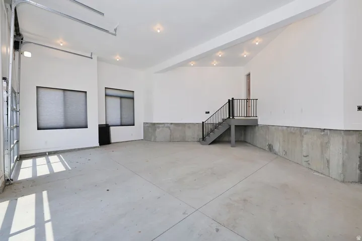 Spacious garage featuring a concrete floor, high vaulted ceilings with recessed lighting, an overhead door with frosted glass panels, two windows with blinds, and a built-in staircase with a black metal railing