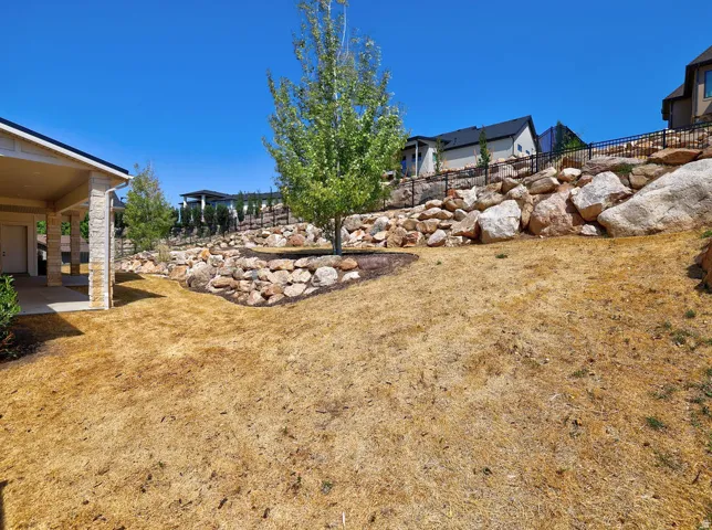 Expansive outdoor space featuring a covered patio with stone columns, a sloped yard, and a rock retaining wall with established trees