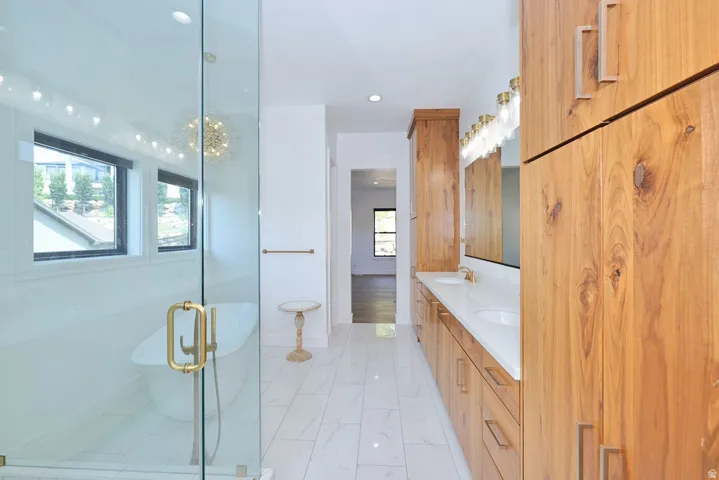 Spacious bathroom featuring a frameless glass shower enclosure with a freestanding soaking tub, dual vanity with undermount sinks and quartz countertops, extensive wood cabinetry, and polished gold-tone hardware throughout