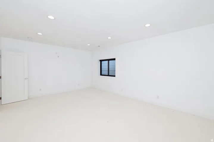 Spacious room featuring plush carpeting, crisp white walls, recessed ceiling lighting, a contemporary white door, and a black-framed window with horizontal blinds