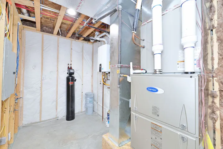 Utility room featuring a Carrier furnace, hot water heater, water softener, exposed ceiling joists, and a visible electrical panel