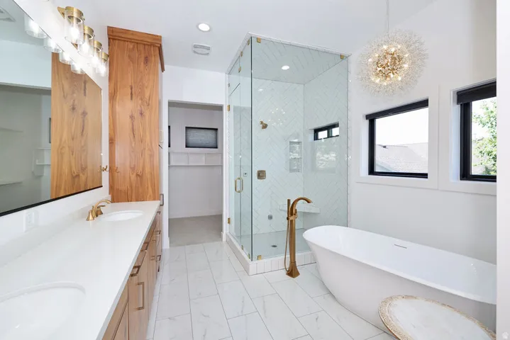 Bathroom featuring a freestanding tub with a floor-mounted brass faucet, a glass-enclosed shower with herringbone tile, a double vanity with light wood cabinetry and white countertops, and white tile flooring