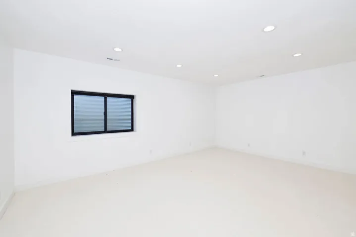 Spacious room featuring neutral carpeting, white walls, recessed lighting, and a dark-framed window with a horizontal blind