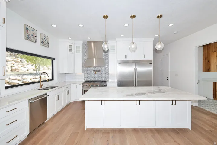 Bright kitchen featuring wood-finish flooring, white cabinetry, substantial island with a stone countertop, stainless steel appliances, and a tiled backsplash