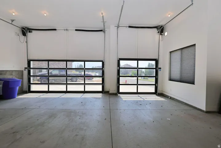 Spacious garage featuring two full-view garage doors with black frames, concrete flooring, recessed ceiling lighting, and a large window with privacy blinds
