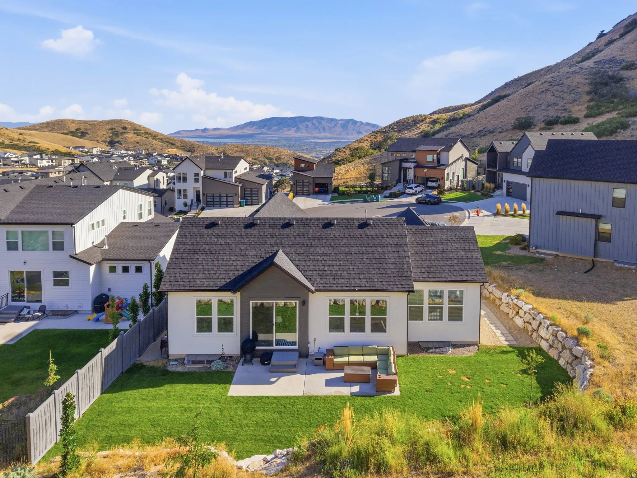 Rear elevation featuring a patio with direct access to a manicured lawn, a privacy fence, multiple windows, and a dark shingle roof