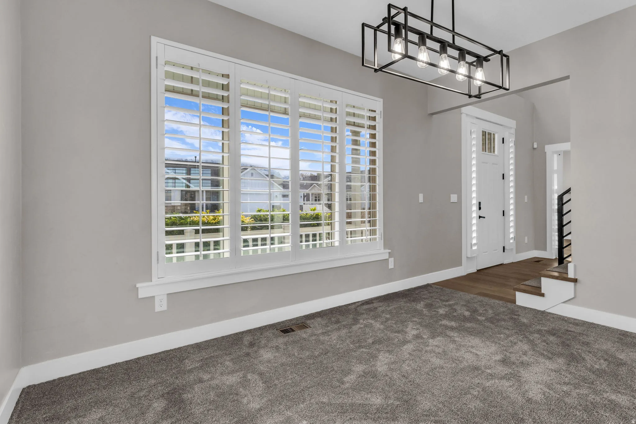 Window with white plantation shutters, gray carpeting, neutral wall paint, and a modern rectangular chandelier