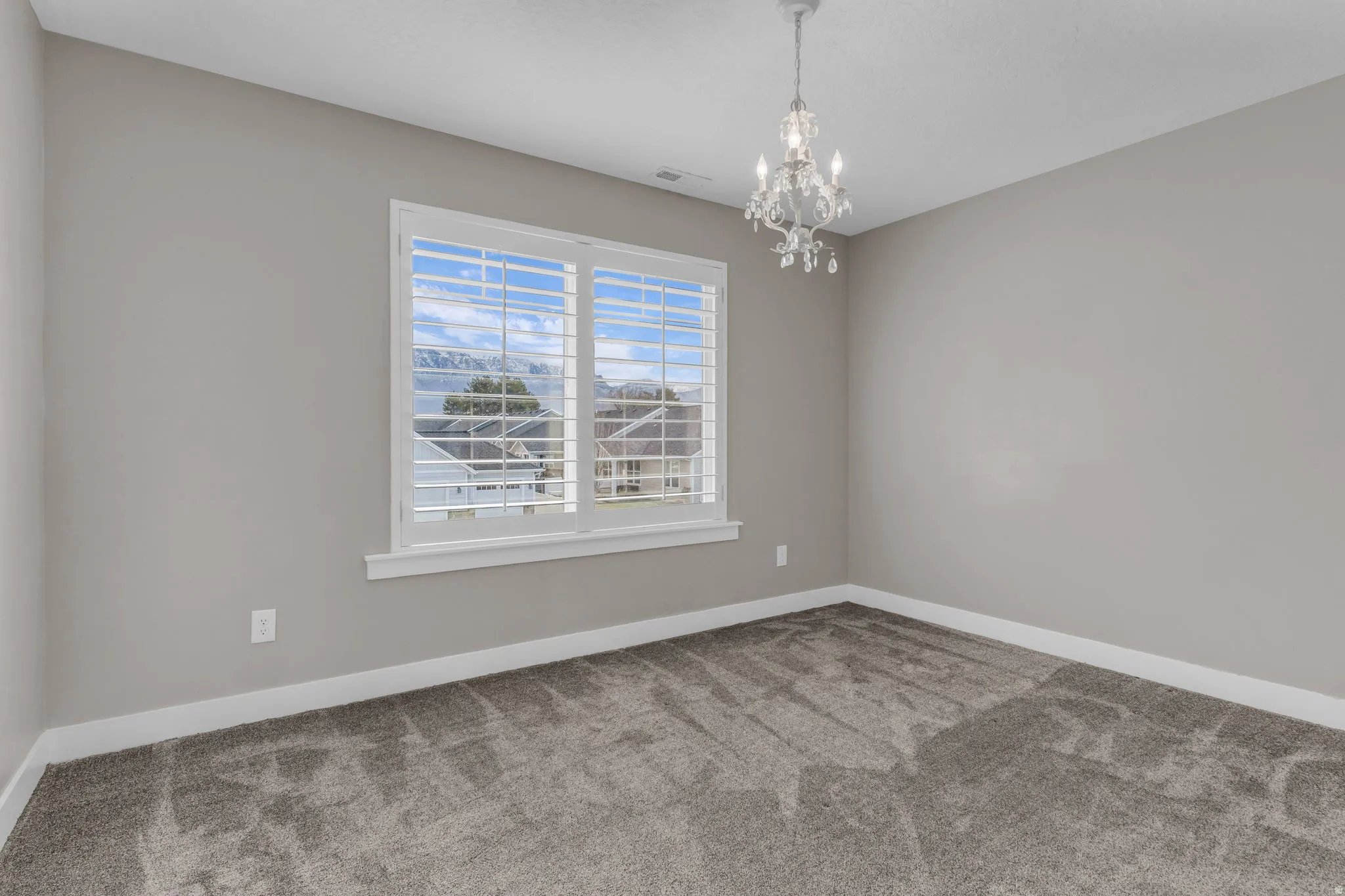 Neutral tone walls with white trim, gray patterned carpet, a crystal chandelier, and a window with white plantation shutters