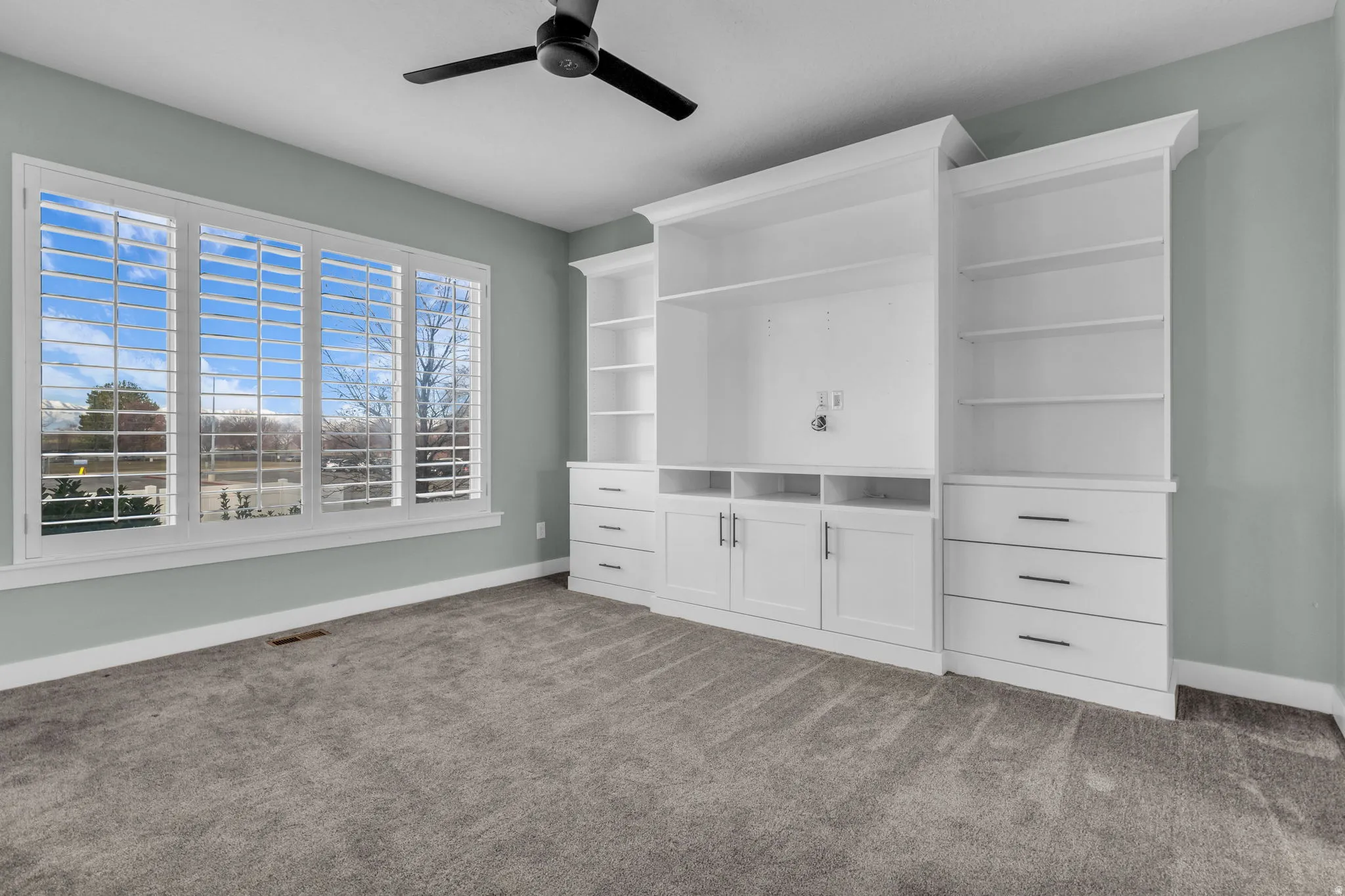 Spacious room featuring a multi-pane window with plantation shutters, custom built-in cabinetry, integrated shelving, and soft gray carpet flooring