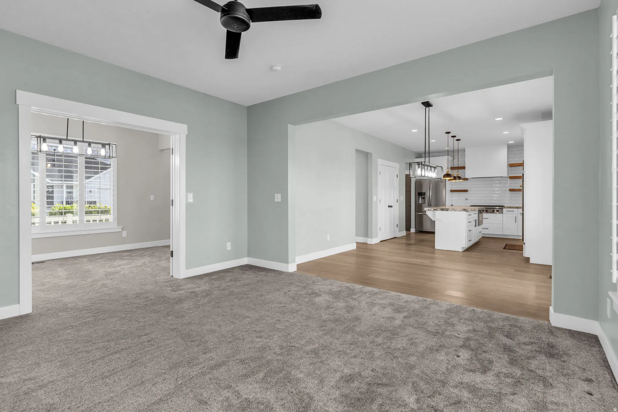 Expansive living area with gray carpeting and a black ceiling fan, opening to a separate room with a large window and a linear light fixture