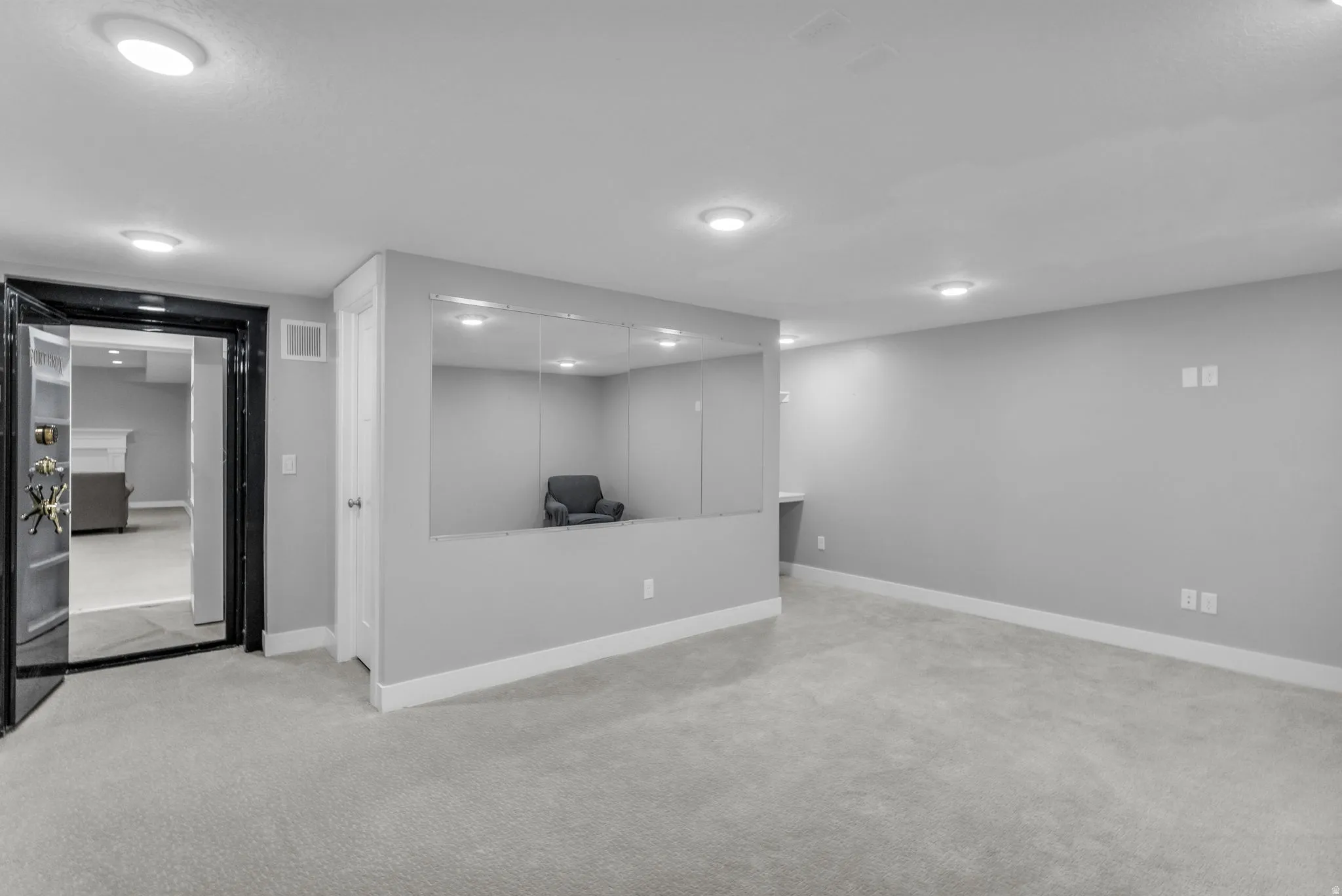 Spacious room with light gray carpeting, recessed lighting, and a prominent dark-framed doorway featuring a vault-style entry