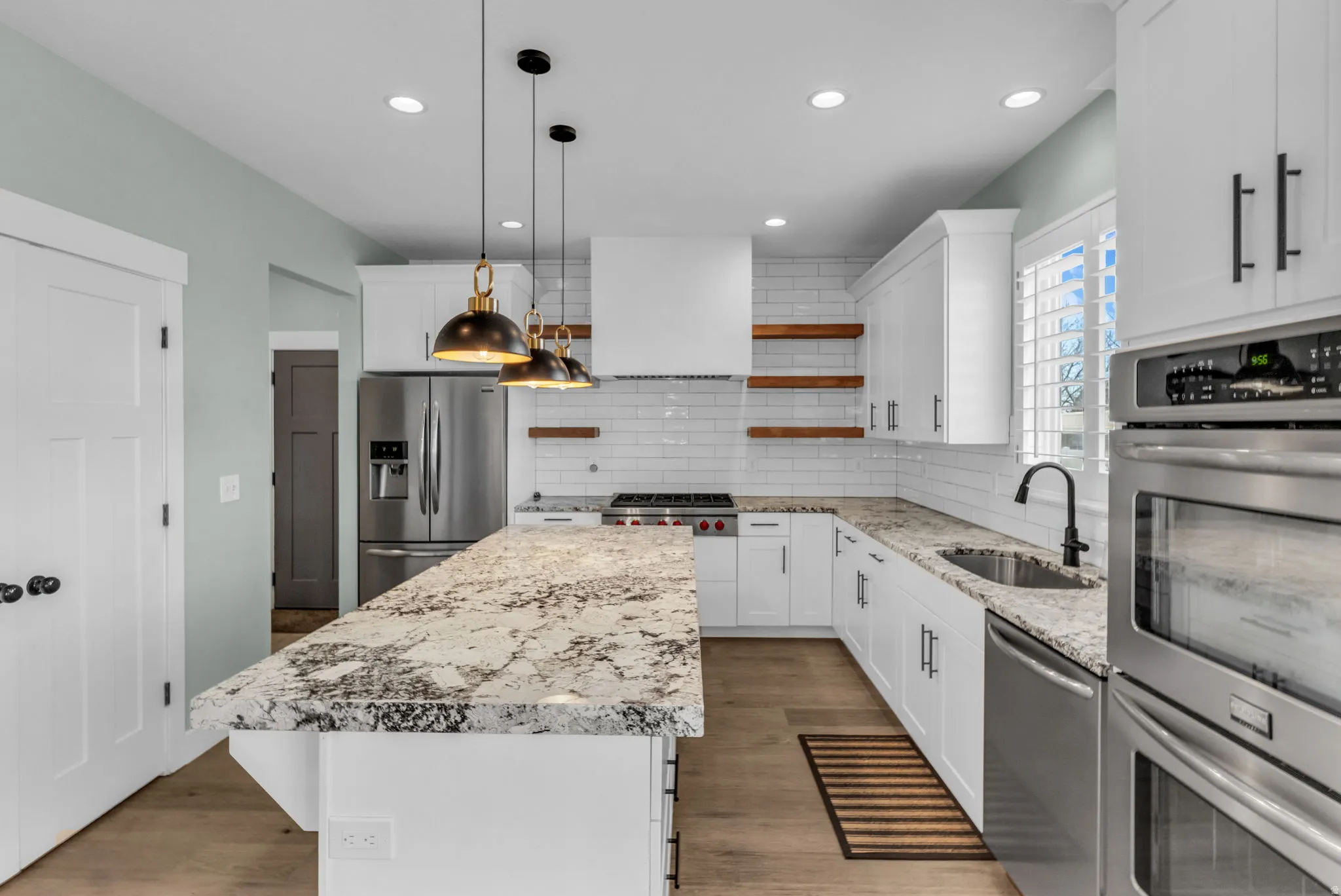 Contemporary kitchen featuring a large island with a substantial stone countertop, white shaker-style cabinetry, stainless steel appliances, a subway tile backsplash, and wood-finish flooring