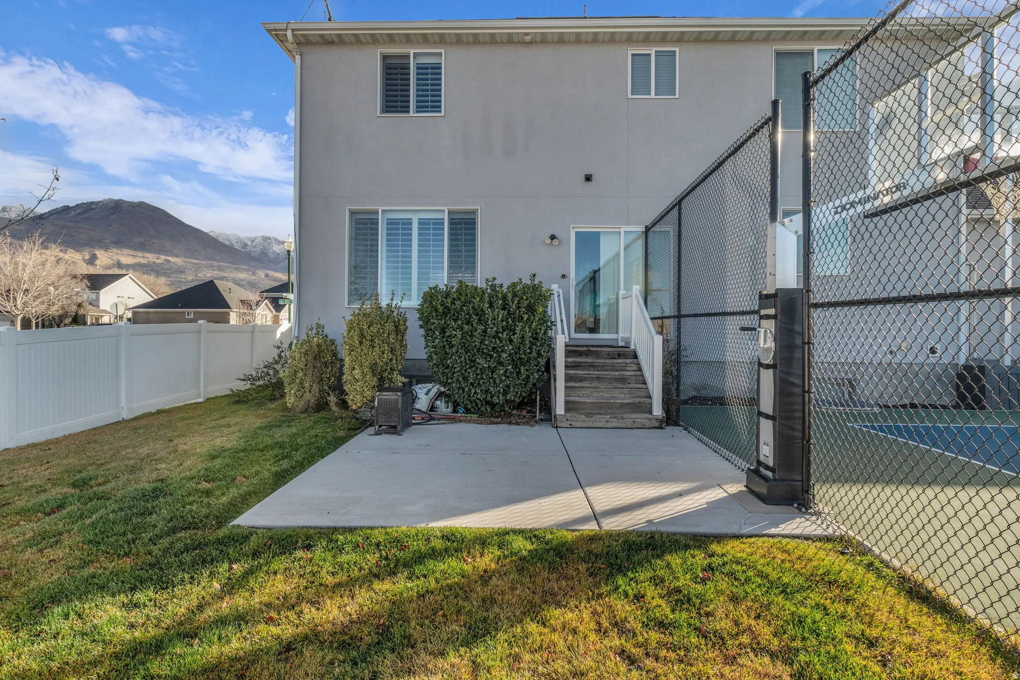 Expansive backyard featuring a concrete patio, mature landscaping, and a chain-link fence enclosing a recreational court