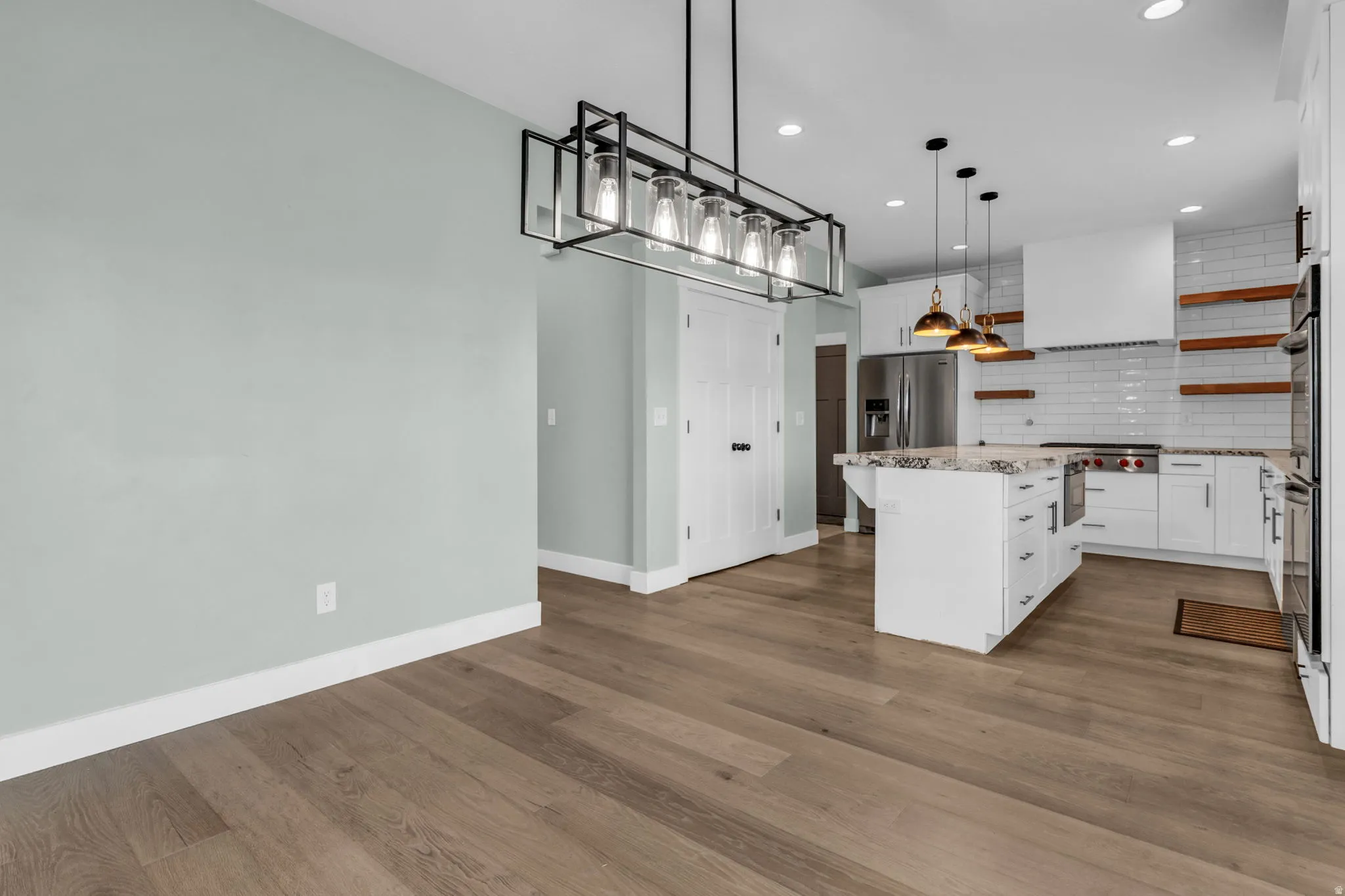 Open-concept kitchen and dining area featuring wood-finish flooring, a contemporary linear chandelier, white shaker cabinetry, a central island with a stone countertop, and stainless steel appliances