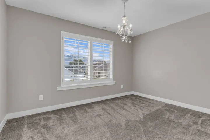 Neutral tone walls with white trim, gray patterned carpet, a crystal chandelier, and a window with white plantation shutters