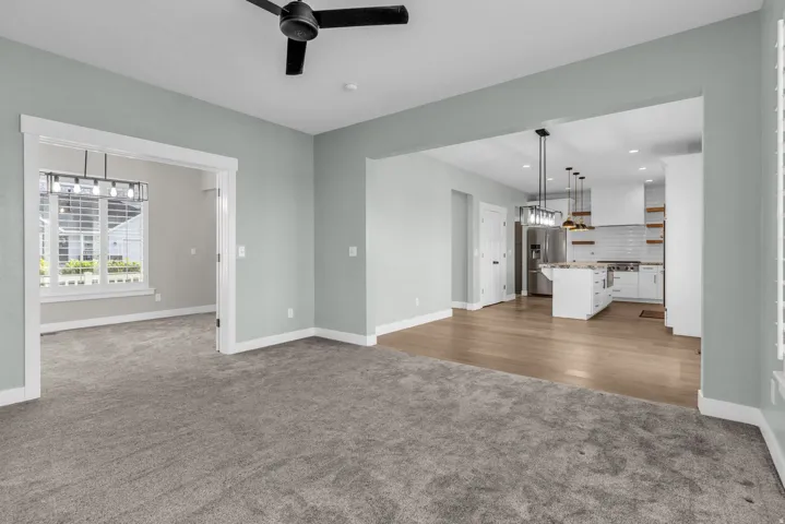 Expansive living area with gray carpeting and a black ceiling fan, opening to a separate room with a large window and a linear light fixture