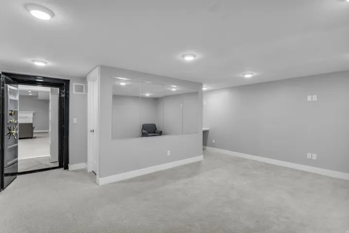 Spacious room with light gray carpeting, recessed lighting, and a prominent dark-framed doorway featuring a vault-style entry