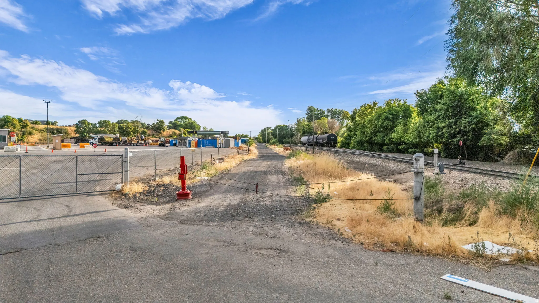 Paved road entrance to a fenced industrial yard with adjacent railway tracks