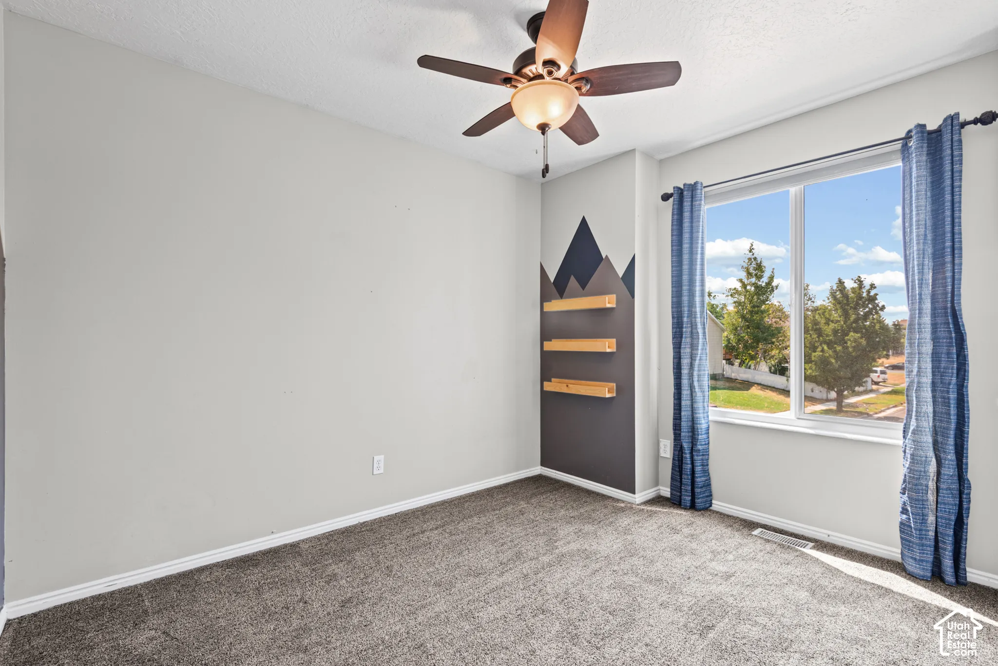 Carpeted flooring, ceiling fan with integrated lighting, window with double pane glass, wall-mounted floating shelves, and a decorative accent wall