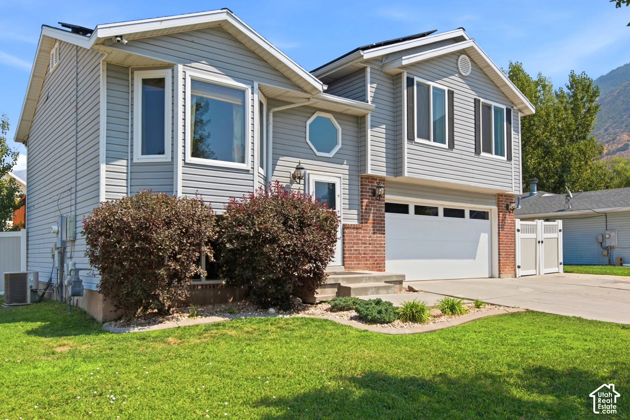Gray siding exterior with a red brick accent, a two-car garage, a concrete driveway, and mature landscaping