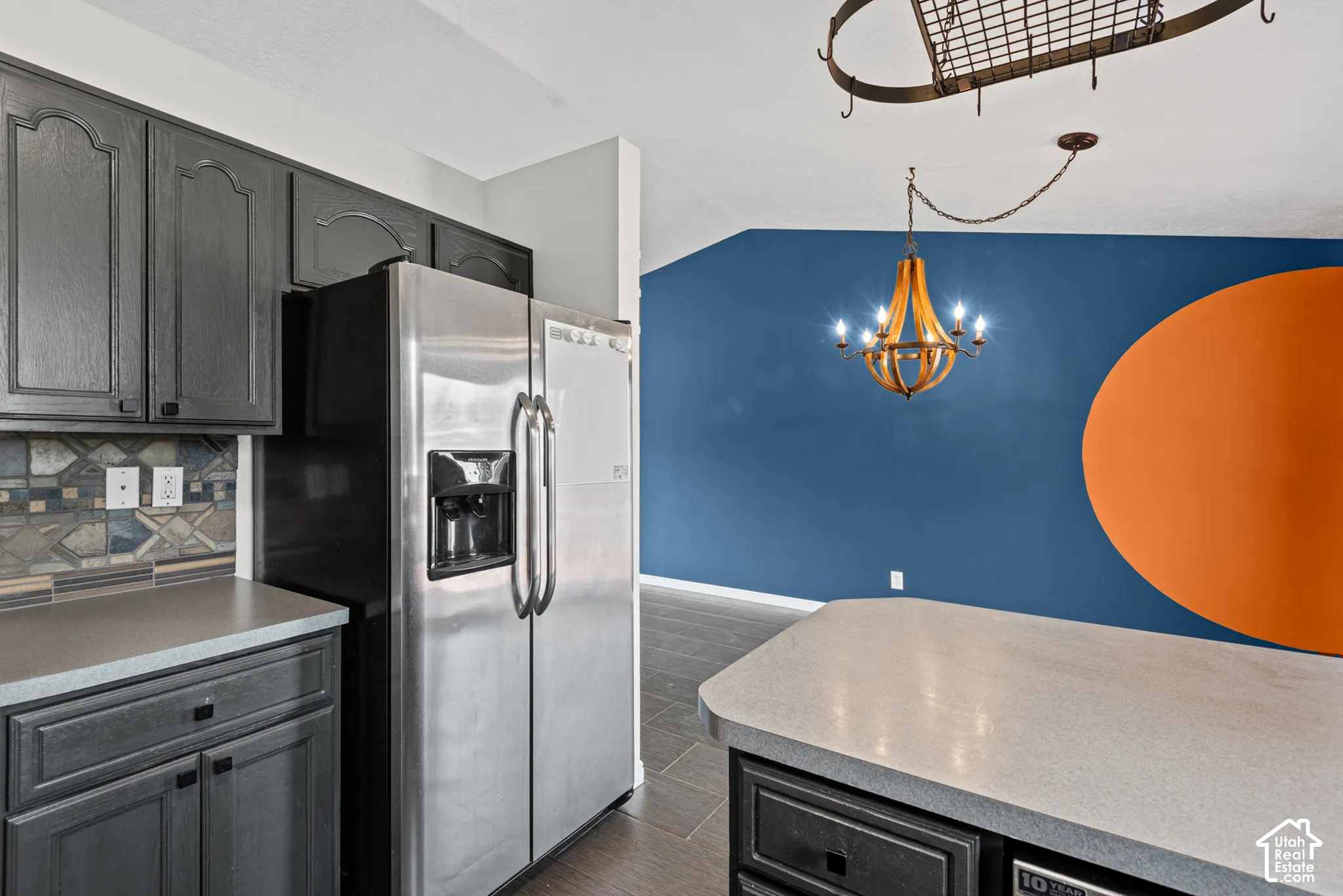 Stainless steel refrigerator, gray cabinetry, patterned tile backsplash, light-toned countertops, and a decorative chandelier