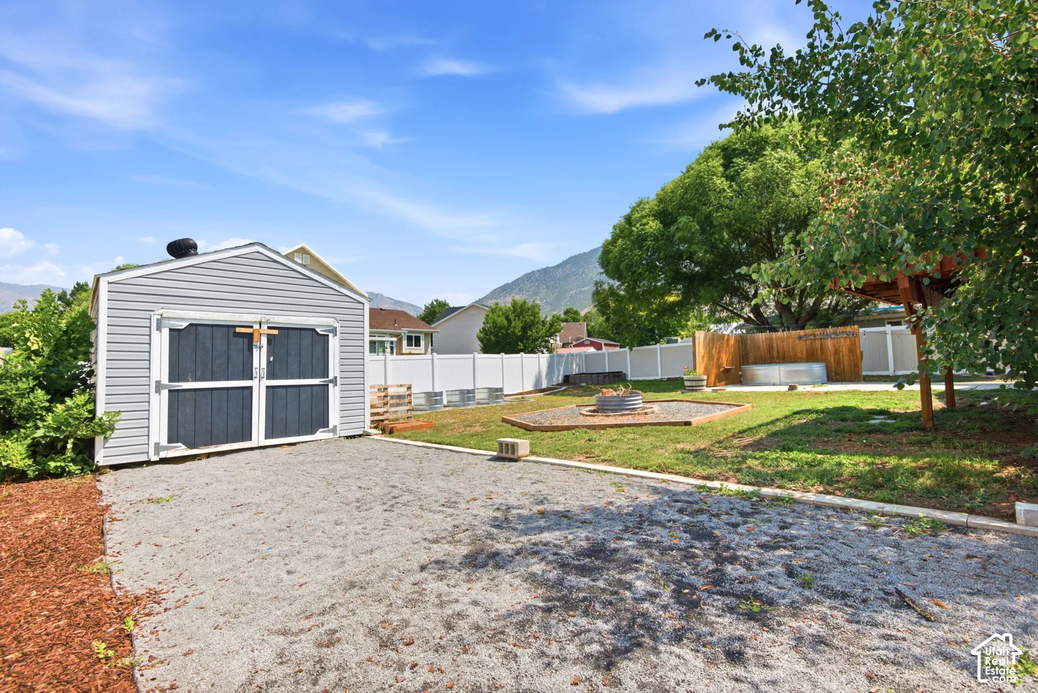 Exterior storage shed with double doors, gravel area, and fenced yard featuring a fire pit and mature trees