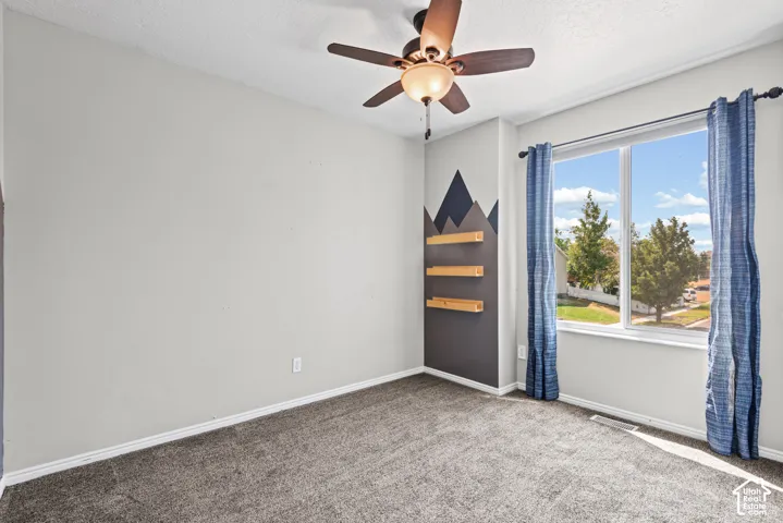 Carpeted flooring, ceiling fan with integrated lighting, window with double pane glass, wall-mounted floating shelves, and a decorative accent wall