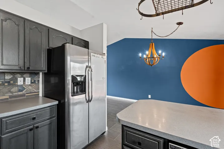 Stainless steel refrigerator, gray cabinetry, patterned tile backsplash, light-toned countertops, and a decorative chandelier