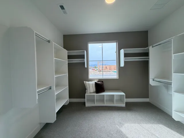 Spacious walk-in closet featuring built-in white shelving, integrated hanging rods, a window, and light gray carpet flooring