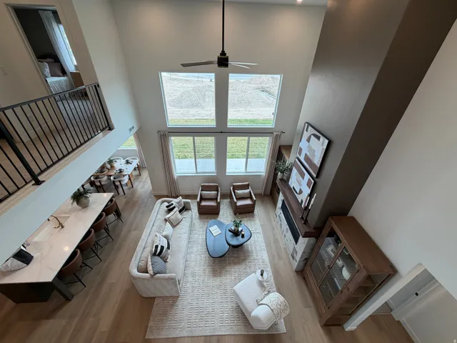 Two-story living area featuring a vaulted ceiling with a contemporary ceiling fan, large windows with exterior views, wood-finish flooring, a stone-clad fireplace with a wood mantle, and an open-concept kitchen island with a light-toned countertop and sin